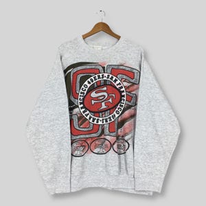 Puede incluir: Sudadera gris claro con el logotipo de los 49ers de San Francisco en rojo y blanco. El logotipo incluye el nombre del equipo y el emblema "SF". La sudadera tiene mangas largas y cuello redondo.