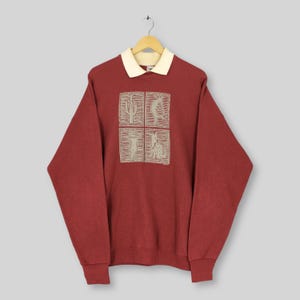 Peut inclure: Un sweat-shirt bordeaux avec un col crème et un motif carré avec des illustrations sur le thème du désert. Le motif comprend un cactus, une personne, une silhouette et un animal.