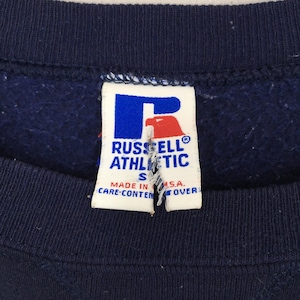 May include: Close-up of a navy blue sweatshirt with a white tag. The tag features the "RUSSELL ATHLETIC" logo in blue and red, with the size "S" and text "MADE IN U.S.A."