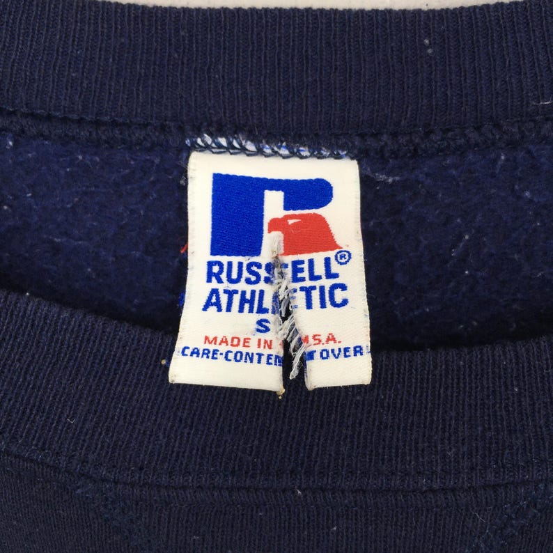 May include: Close-up of a navy blue sweatshirt with a white tag. The tag features the "RUSSELL ATHLETIC" logo in blue and red, with the size "S" and text "MADE IN U.S.A."