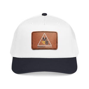 May include: A white baseball cap with a navy blue brim and a black button on top. The cap features a brown leather patch with the text "Mindful Barber" and a logo with the letters "M" and "B" inside a triangle.