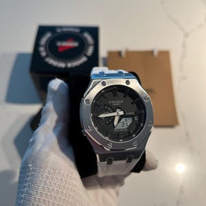 May include: A Casio G-Shock watch featuring a silver-toned octagonal metal bezel with eight visible screws, a black analog-digital dial with white hands, and a white silicone strap. The watch face displays the G-Shock Casio logo and digital time.
