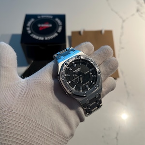 May include: A silver-toned G-Shock Casio wristwatch featuring an octagonal bezel with exposed screws, a black analog-digital dial, and a matching metallic link bracelet. The watch is presented with its black branded box and a brown paper bag.
