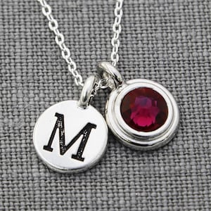 May include: A silver necklace with two charms. One charm is a round disc with the letter "M" engraved on it. The other charm is a round disc with a red gemstone in the center.