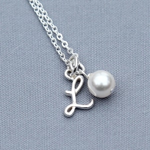 Puede incluir: Un collar de plata con un dije de letra L en cursiva y un pequeño colgante de perla blanca.