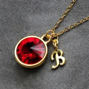 May include: A gold-colored necklace with a large, round, faceted red gemstone pendant. A small gold letter "B" charm hangs next to the pendant. The necklace chain is delicate and gold, set against a gray background.