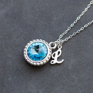 Op de afbeelding: Een zilveren ketting met een lichtblauwe kristallen hanger en een zilveren bedel met de letter 'L'.