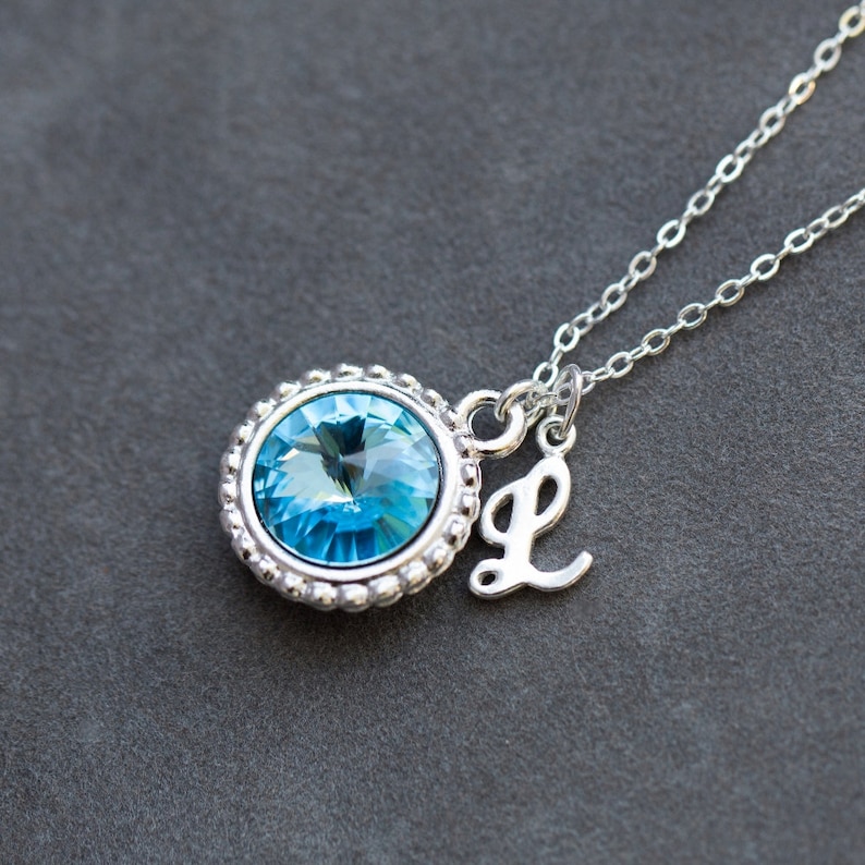 Op de afbeelding: Een zilveren ketting met een lichtblauwe kristallen hanger en een zilveren bedel met de letter 'L'.