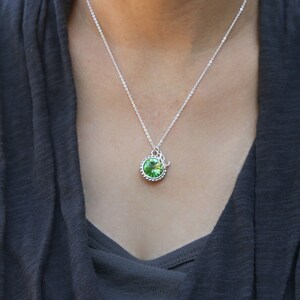 Op de afbeelding: Een zilveren ketting met een groene kristallen hanger. De hanger is rond en heeft een zilveren rand.