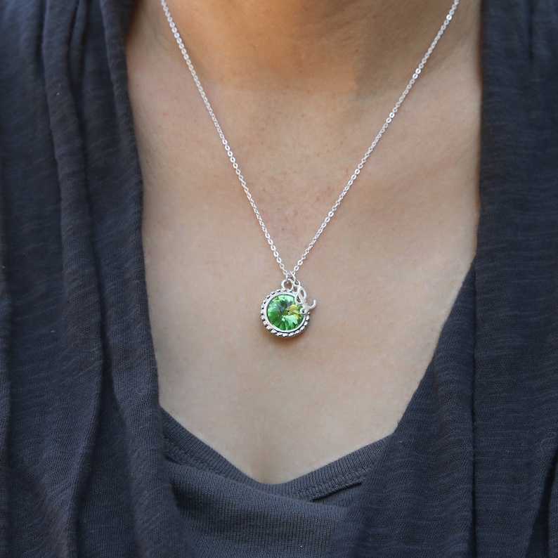 Op de afbeelding: Een zilveren ketting met een groene kristallen hanger. De hanger is rond en heeft een zilveren rand.