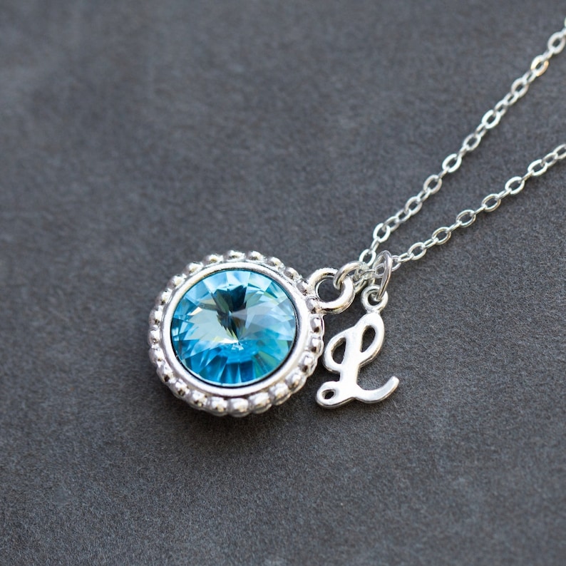 Op de afbeelding: Een zilveren ketting met een blauwe kristallen hanger en een zilveren bedel met de letter L.