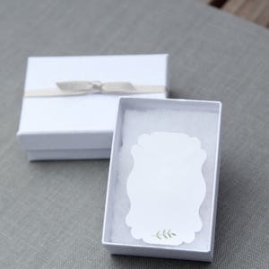 Op de afbeelding: Witte cadeaudoos met een wit lint en strik. De doos is open en bevat een witte kaart met een groen bladerenontwerp.
