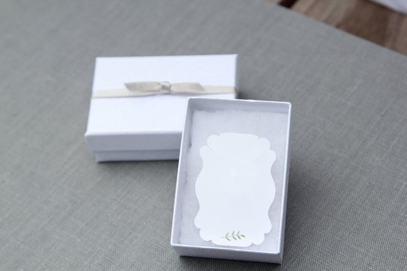 Op de afbeelding: Witte cadeaudoos met een wit lint en strik. De doos is open en bevat een witte kaart met een groen bladerenontwerp.