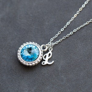 Op de afbeelding: Een zilveren ketting met een blauwe kristallen hanger en een zilveren bedel met de letter L.