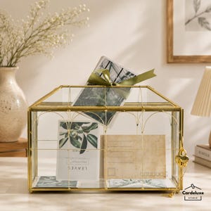May include: A gold-colored, glass and metal decorative box with a house-like shape. The box contains postcards and a card with the word "LEAVES". A green ribbon is tied around a postcard. A gold heart-shaped lock and key are on the side.