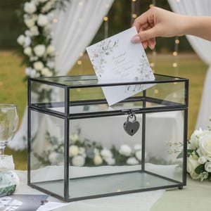 May include: A clear glass card box with a black metal frame and a heart-shaped lock. A person is inserting a card into the box. The card has floral designs and text. The box is on a table with flowers and a glass.
