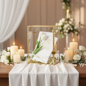 May include: A gold-framed glass box displays a card with the words "His Vows" and a white tulip. The box sits on a white tablecloth, surrounded by lit candles and floral arrangements, creating a romantic ambiance.