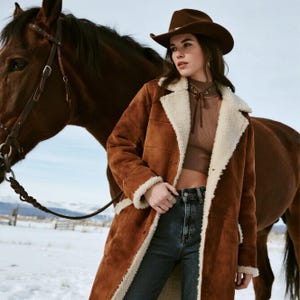 Könnte beinhalten: Ein brauner Wildledermantel mit cremefarbenem Shearling-Futter und -Besatz. Der Mantel wird mit einem braunen Cowboyhut und dunkelblauen Jeans getragen. Im Hintergrund steht ein braunes Pferd in einer verschneiten Landschaft.