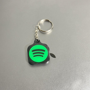 Può includere: Un portachiavi nero e verde con il logo Spotify. Il portachiavi presenta un'etichetta rettangolare nera con un cerchio verde contenente tre linee orizzontali, il logo Spotify. È attaccato a un portachiavi argentato.