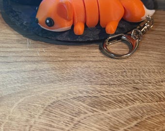 Peut inclure: Un porte-clés renard orange imprimé en 3D avec un fermoir argenté. Le renard a des parties du corps segmentées et une queue à bout blanc. Le renard est posé sur une surface en bois.