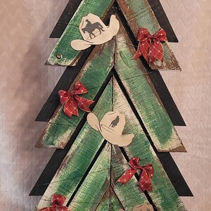 May include: Wooden Christmas tree with a rustic design. The tree is made of green and brown planks, decorated with cowboy hat cutouts, red bows, and a large bow at the top. The base reads "Merry Christmas, Y'all!"