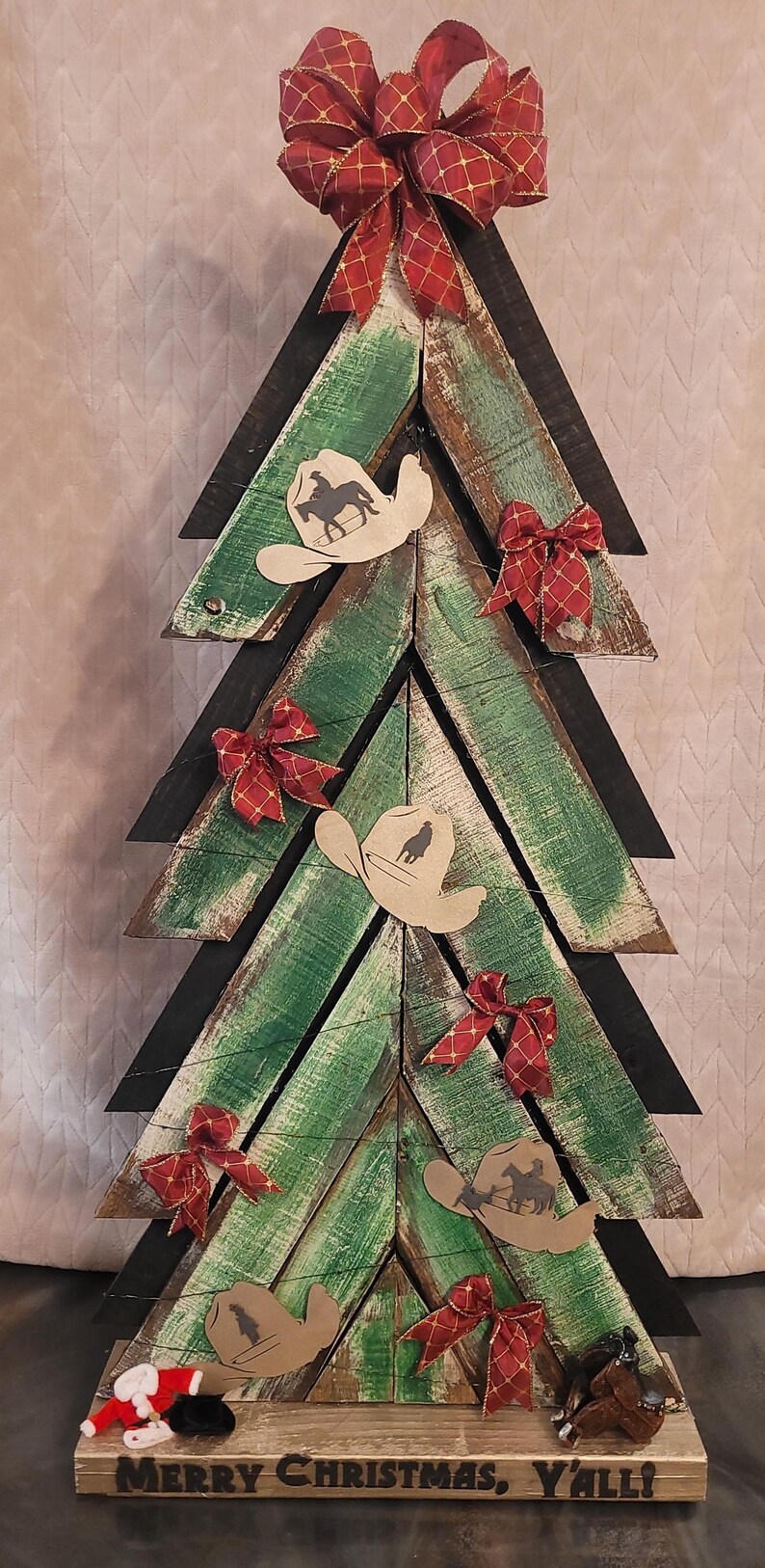 May include: Wooden Christmas tree with a rustic design. The tree is made of green and brown planks, decorated with cowboy hat cutouts, red bows, and a large bow at the top. The base reads "Merry Christmas, Y'all!"