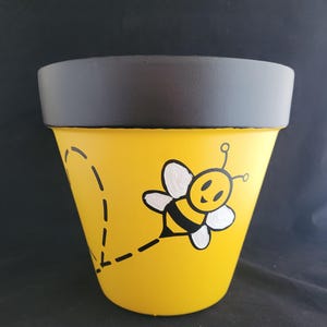 May include: A yellow flower pot with a black rim, decorated with a cartoon bee. The bee has white wings and a black and yellow body. A dotted line shows the bee's flight path.