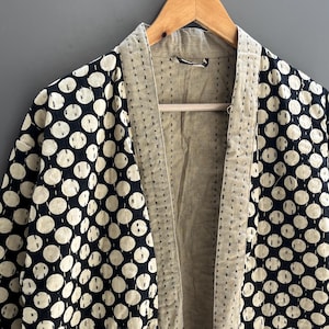 May include: A patterned kimono-style jacket with a cream-coloured interior and a black and cream polka dot exterior. The jacket is hanging on a wooden hanger against a grey background. The edges are stitched.