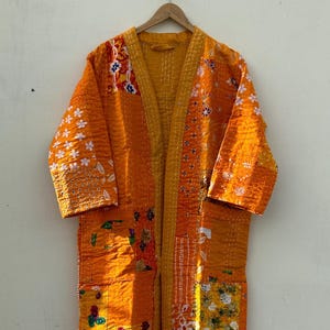 Peut inclure: Robe kimono orange en patchwork avec manches trois-quarts. Motifs floraux et géométriques en orange, jaune et blanc. Suspendue à un cintre en bois sur fond blanc.