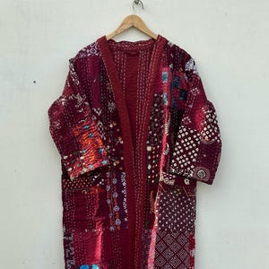 Könnte beinhalten: Langer, burgunderfarbener Patchwork-Bademantel mit einer Vielzahl von Mustern und Texturen. Der Bademantel besteht aus verschiedenen Stoffstücken, die zusammengenäht wurden, wodurch ein einzigartiges und farbenfrohes Design entsteht. Der Bademantel hängt an einem Holzbügel.