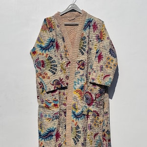 May include: A cream-coloured quilted robe with a vibrant floral pattern in shades of blue, yellow, pink, and green. The robe features a light pink trim and a matching belt. It is hanging on a metal hanger.