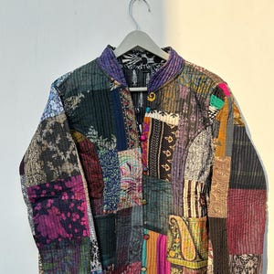 Peut inclure: Veste patchwork avec un col officier, présentant une variété de motifs et de couleurs de tissus. La veste est suspendue à un cintre en bois. La palette de couleurs comprend des nuances de violet, de vert, de rose et d'or.