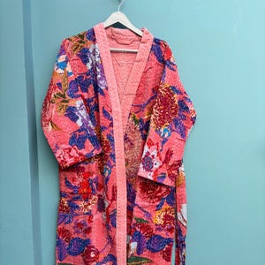 Peut inclure: Une longue robe à motifs floraux dans les tons de rose, violet, bleu et rouge est suspendue à un cintre blanc. La robe présente une bordure rose contrastante et une ceinture assortie. Le tissu semble être matelassé.