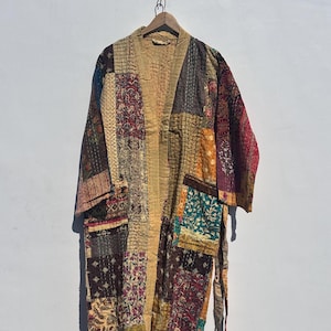 Könnte beinhalten: Ein Patchwork-Kimono-Roben mit einer Vielzahl von gemusterten Stoffquadraten in Braun-, Beige-, Rot- und Grüntönen. Die Robe hat lange Ärmel und einen Bindegürtel. Das Kleidungsstück hängt an einem Holzbügel vor einer weißen Wand.