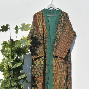 May include: A long, open-front robe with a green interior and trim, and a patterned exterior in shades of green, orange, and yellow. The robe is hanging on a wooden hanger against a white wall. The fabric appears to be quilted.