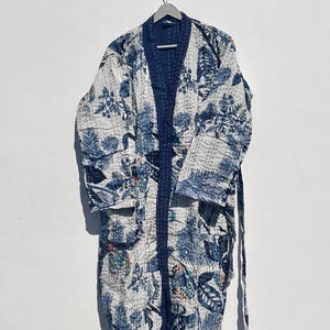May include: A white and blue floral patterned robe with a dark blue trim and belt. The robe is hanging on a hanger against a white background. The floral pattern includes leaves and other botanical elements.
