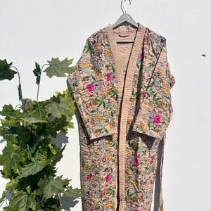 May include: A long, floral-patterned robe in shades of pink, green, yellow, and purple hangs on a grey hanger. The robe has a light pink trim and a matching belt. The background is a plain white wall.