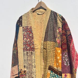 May include: A patchwork kimono-style jacket with a V-neck and long sleeves. The jacket is made of various fabric patches in shades of beige, brown, red, and teal. It hangs from a wooden hanger against a white background.