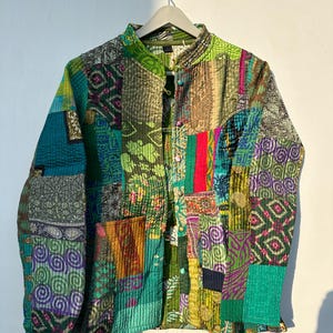 Peut inclure: Veste patchwork aux tons de vert, turquoise, violet et rouge, suspendue à un cintre blanc. La veste est composée de divers morceaux de tissu cousus ensemble, créant un design unique et texturé. Le col est montant et la veste se ferme par des boutons.