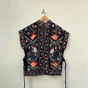 May include: A navy blue embroidered jacket with a mandarin collar and short sleeves. The jacket features a floral design in shades of coral, blue, and yellow. It has a tie closure at the waist and is hanging on a wooden hanger.