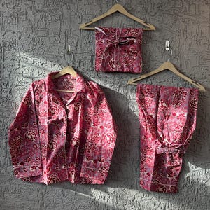 May include: A pink floral pajama set, including a button-down shirt, pants, and a wrapped package, all hanging on wooden hangers against a textured gray wall. The set features a repeating floral pattern.