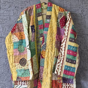 May include: A patchwork jacket in a variety of colors, including yellow, pink, green, and red. The jacket is made of different fabric pieces sewn together, creating a unique and textured look. It hangs on a wooden hanger against a gray wall.