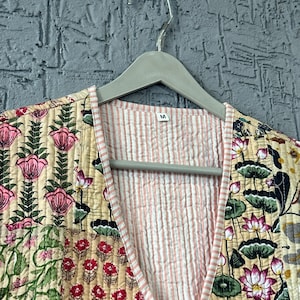 May include: A quilted jacket with a floral pattern in pink, green, and cream. The interior lining is striped in pink and white. The jacket is hanging on a gray hanger against a textured gray wall. The label reads "M".