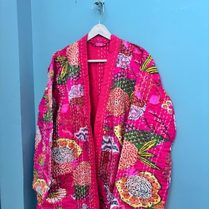 May include: A vibrant pink floral patterned robe hanging on a white hanger. The robe features a quilted design with a variety of colorful floral and leaf patterns. The background is a solid light blue wall.
