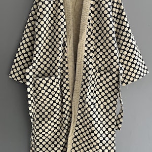 Peut inclure: Robe de style kimono &agrave; motifs de pois noirs et blancs, avec un int&eacute;rieur cr&egrave;me et des poches. La robe est suspendue &agrave; un cintre en bois sur un fond gris.
