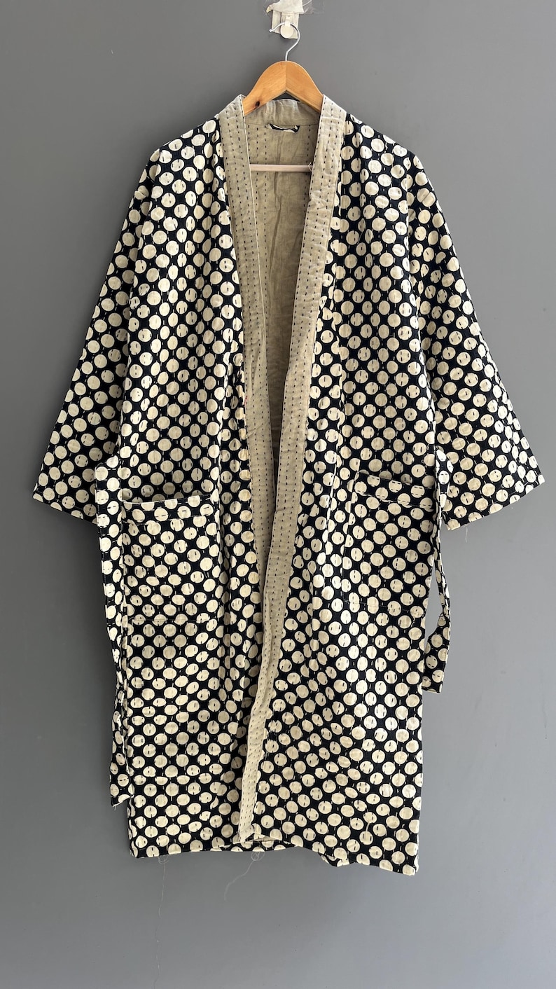 Peut inclure: Robe de style kimono &agrave; motifs de pois noirs et blancs, avec un int&eacute;rieur cr&egrave;me et des poches. La robe est suspendue &agrave; un cintre en bois sur un fond gris.