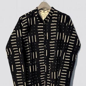 Puede incluir: Chaqueta negra con un patrón de rayas verticales blancas. La chaqueta tiene cuello mandarín, dos bolsillos delanteros y cierre de botones. La chaqueta está colgada en una percha de madera.