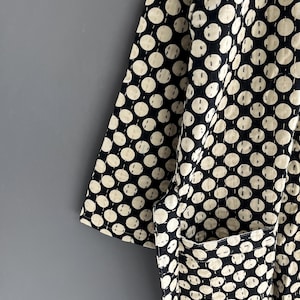 Peut inclure: Gros plan sur un v&ecirc;tement noir et cr&egrave;me &agrave; pois r&eacute;p&eacute;t&eacute;s. Le tissu a un aspect matelass&eacute; avec des coutures visibles. Une poche est visible sur le c&ocirc;t&eacute; droit. Le v&ecirc;tement est probablement une robe de chambre ou une veste.