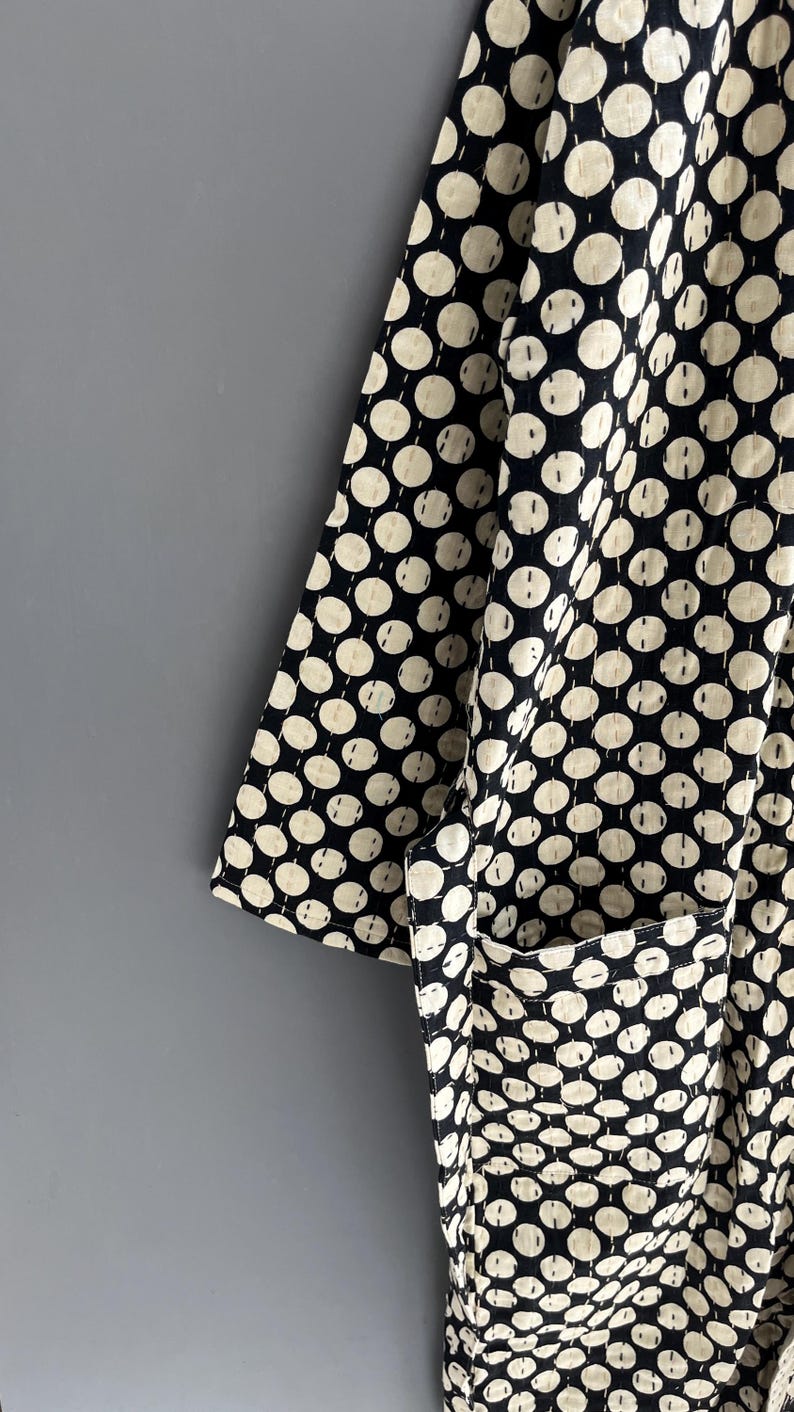 Peut inclure: Gros plan sur un v&ecirc;tement noir et cr&egrave;me &agrave; pois r&eacute;p&eacute;t&eacute;s. Le tissu a un aspect matelass&eacute; avec des coutures visibles. Une poche est visible sur le c&ocirc;t&eacute; droit. Le v&ecirc;tement est probablement une robe de chambre ou une veste.