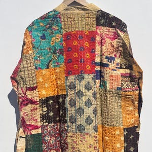 May include: A patchwork jacket with a variety of colorful fabric squares. The jacket features a mix of floral and geometric patterns in shades of teal, red, gold, and brown. The jacket is hanging on a wooden hanger.